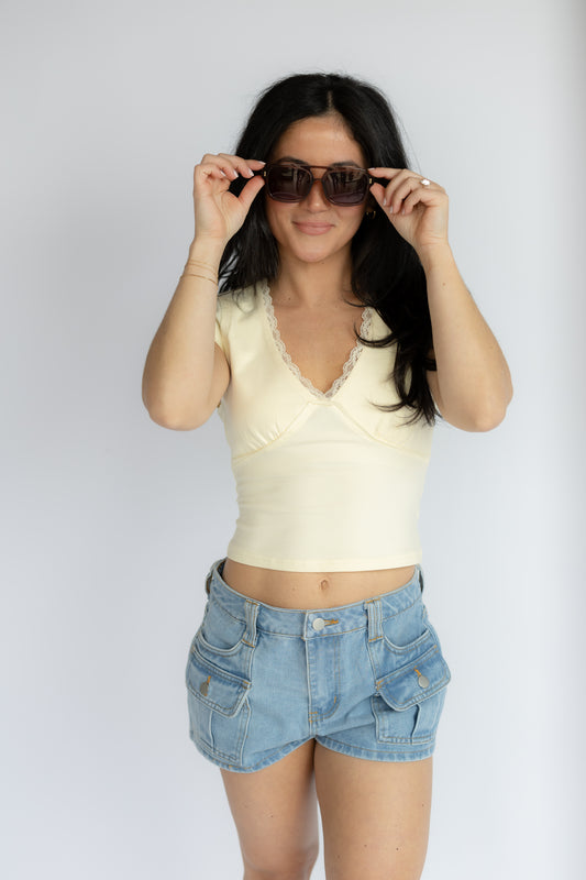 Woman wearing a yellow top and denim shorts, holding sunglasses against a white background