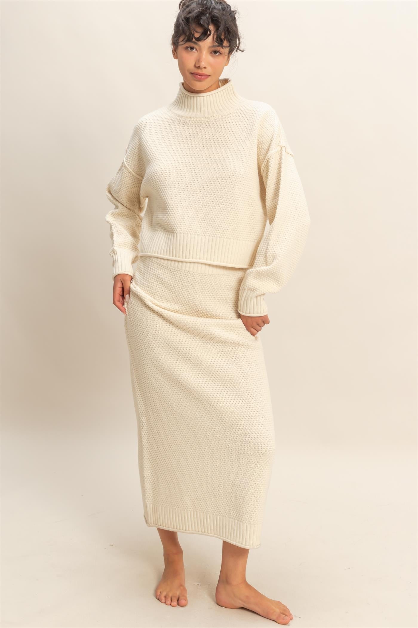 Model wearing ivory textured knit sweater and matching midi skirt set with high neckline and relaxed fit.
