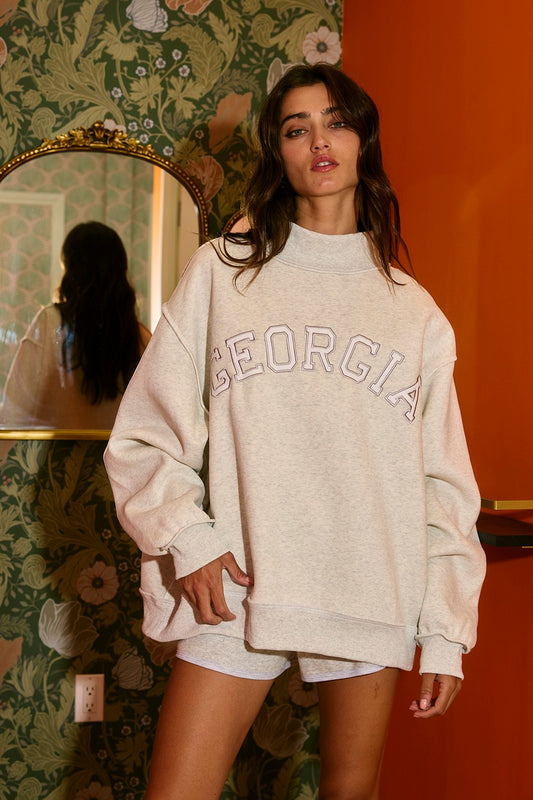 Woman wearing oversized GEORGIA embroidered mock neck sweatshirt and shorts in neutral fleece.