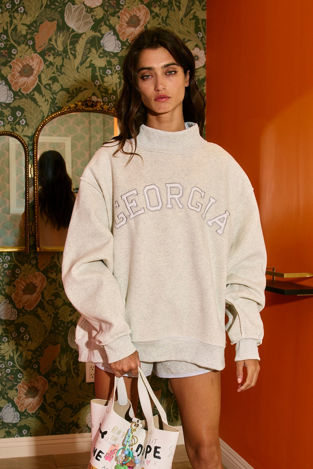 Woman wearing a 'Georgia' sweatshirt in a room with floral wallpaper and an orange wall.