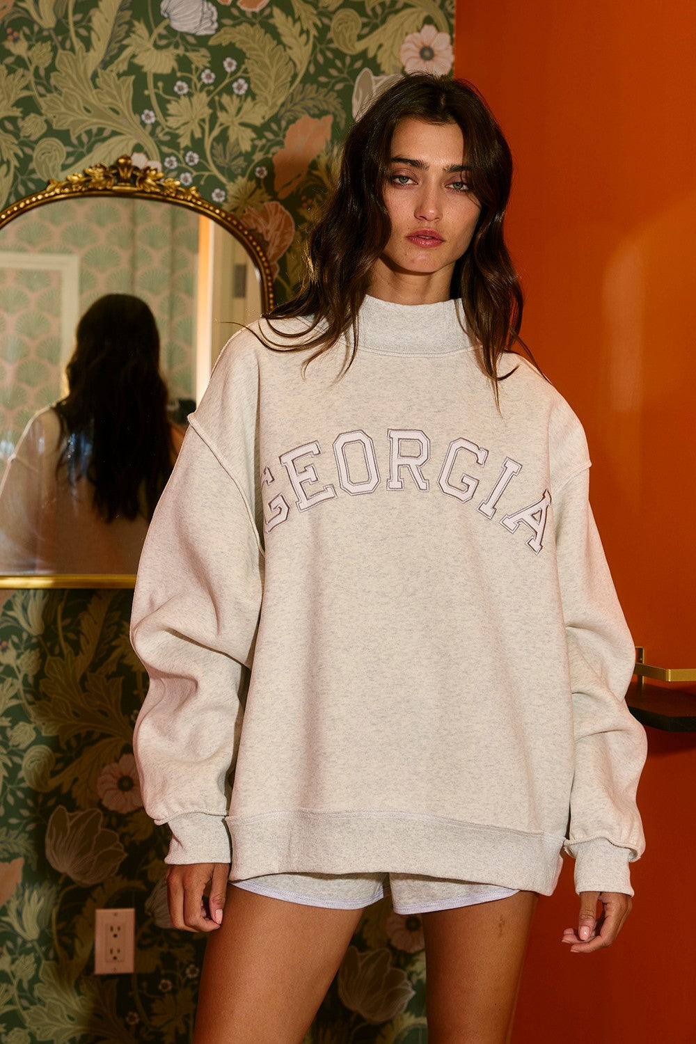 Woman wearing a beige 'Georgia' sweatshirt in a room with floral wallpaper and an orange wall.