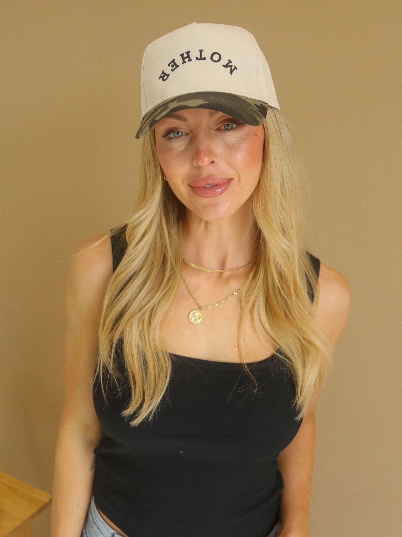 Woman wearing a beige and camo two-tone trucker hat with upside-down embroidered “mother” text styled with a black tank and gold jewelry.