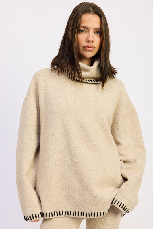 Woman wearing a beige oversized turtleneck sweater with black contrast stitching and matching knit pants.