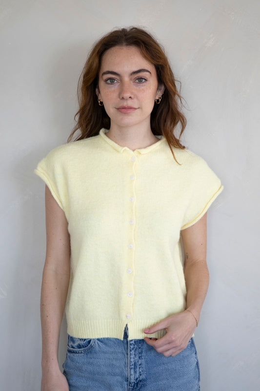Woman wearing a light yellow sleeveless knit vest cardigan with rolled edges and button-up front styled with blue jeans.