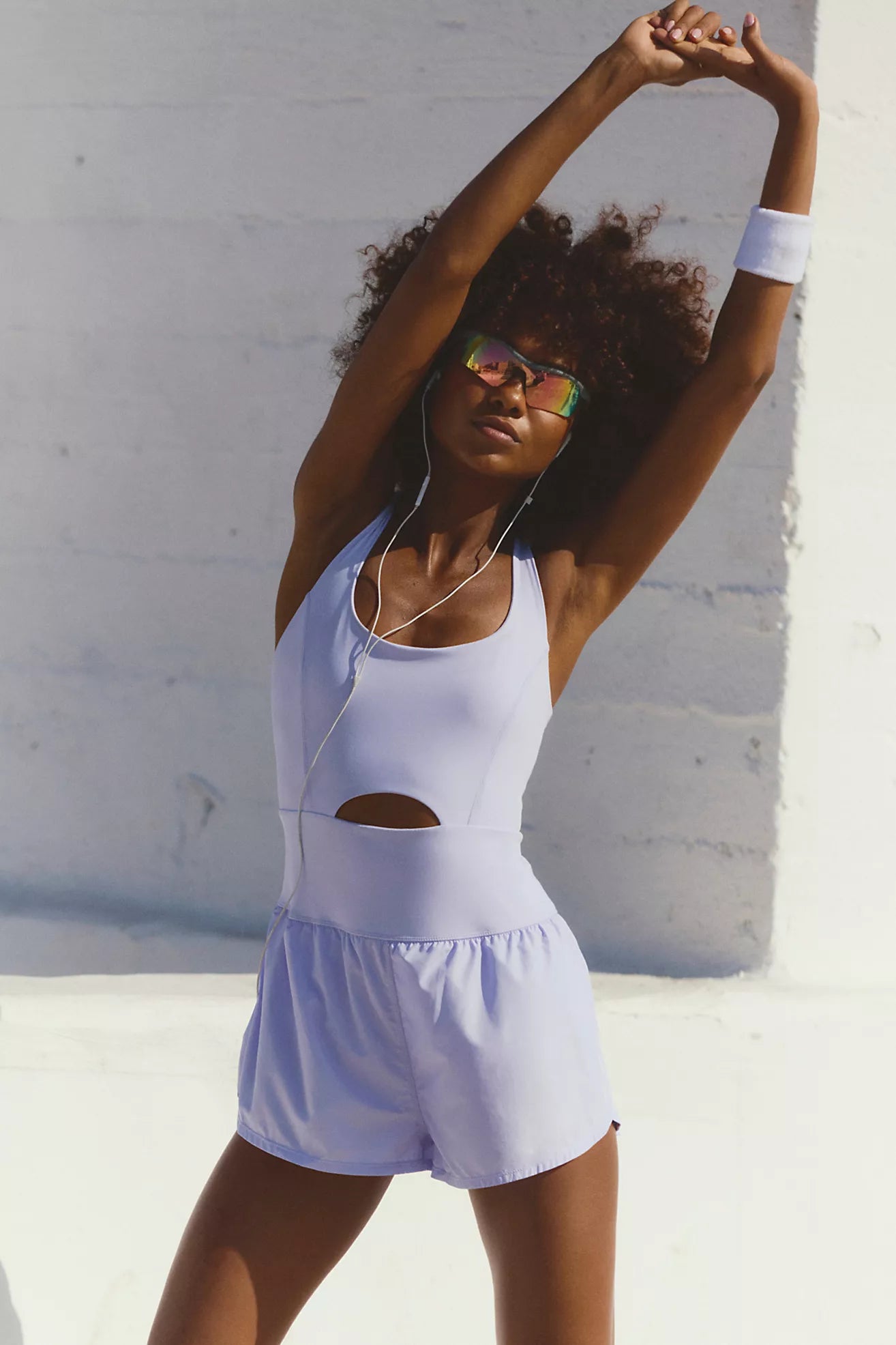 Model stretching outdoors wearing the Righteous Runsie Iris in light purple with cutout detail and retro-style shorts.