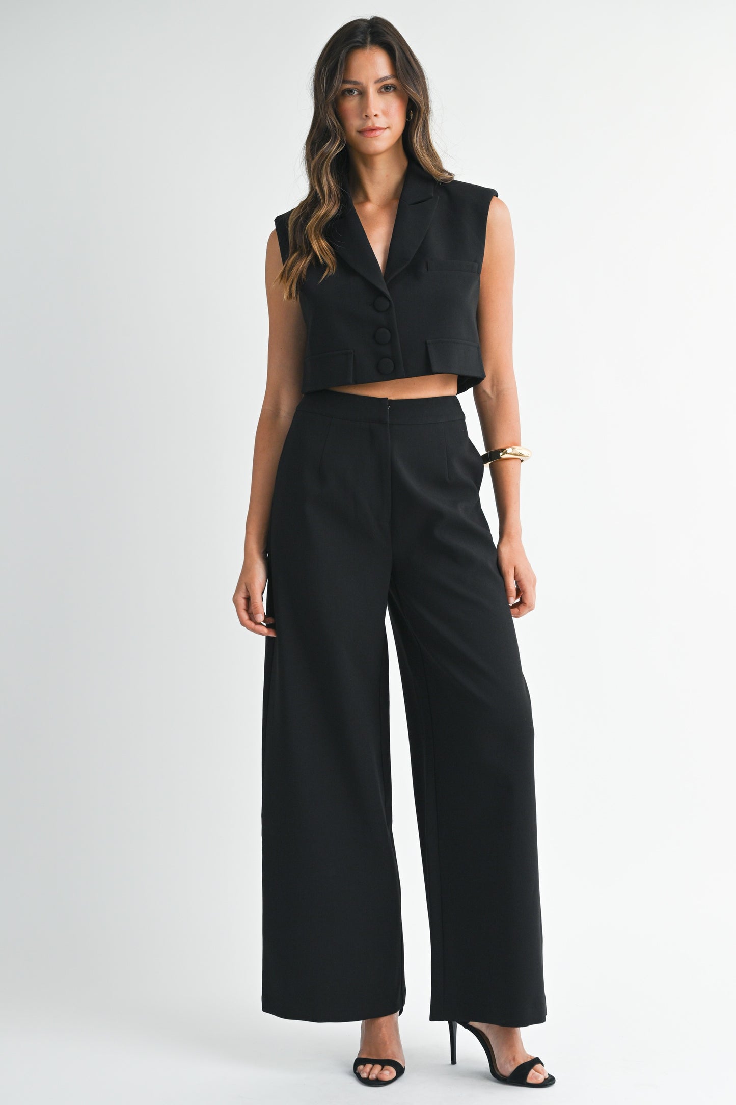 Model wearing a sleeveless black cropped vest and matching wide-leg pants with heels against a white background
