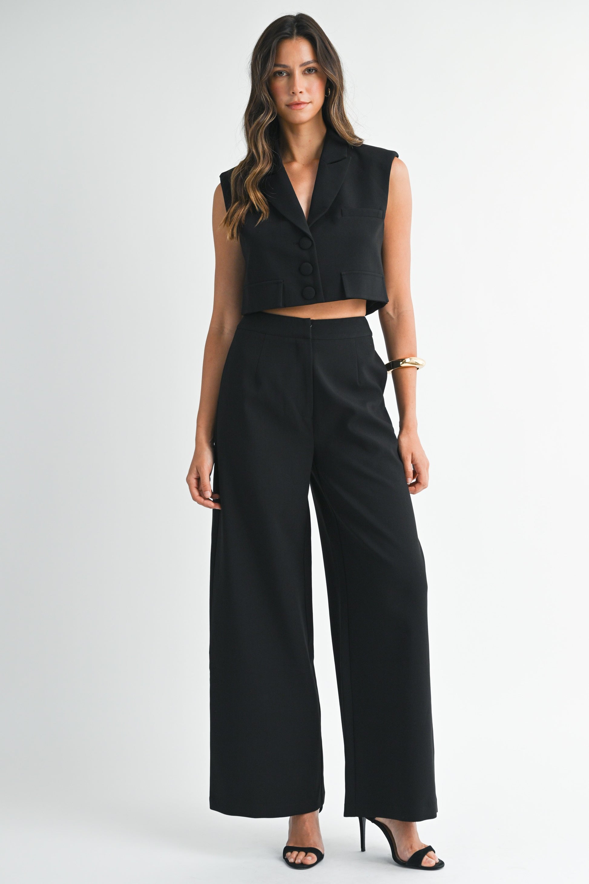 Model wearing a sleeveless black cropped vest and matching wide-leg pants with heels against a white background