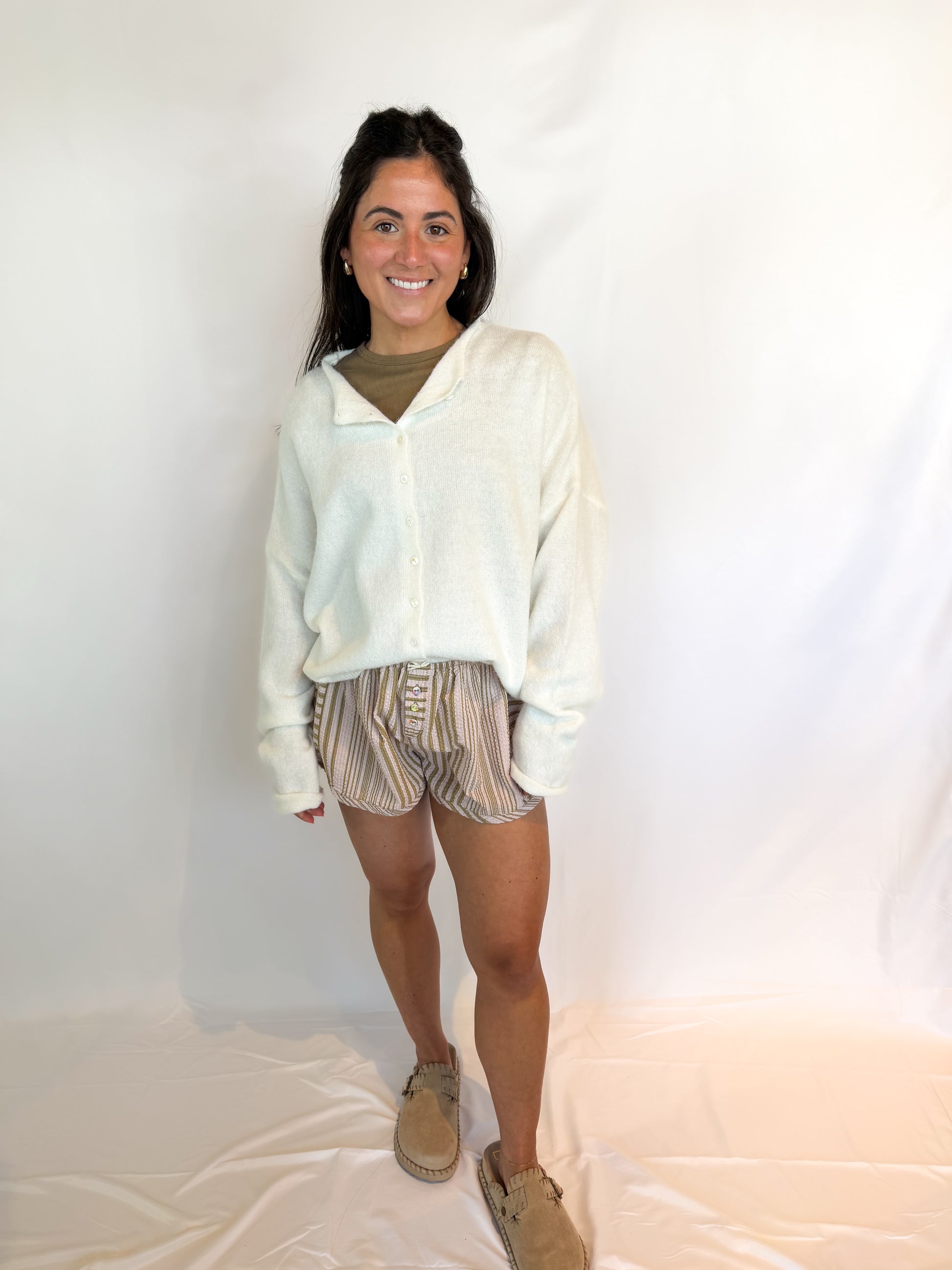 Woman wearing a light-colored cardigan, patterned shorts, and beige shoes against a white background