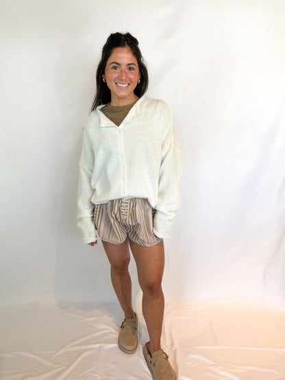Woman wearing a light-colored cardigan, patterned shorts, and beige shoes against a white background