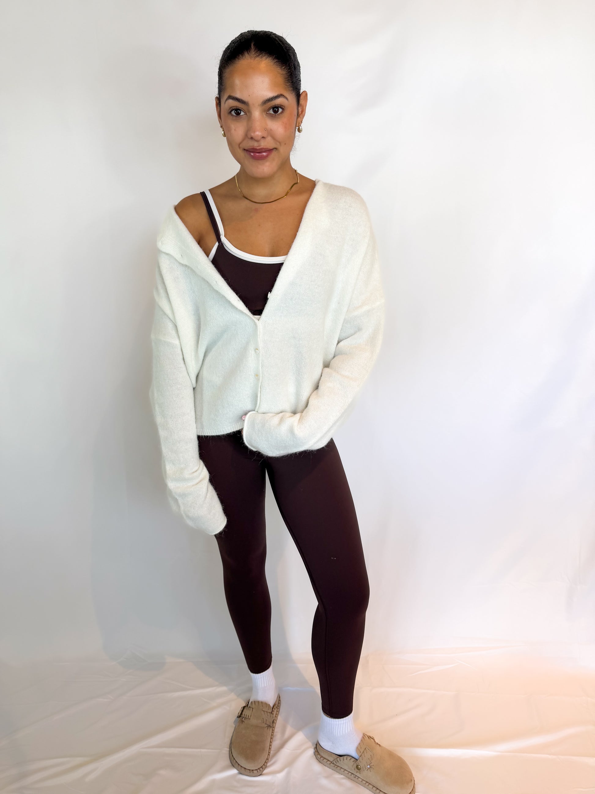 Woman wearing a white cardigan over a dark top with brown pants on a plain background