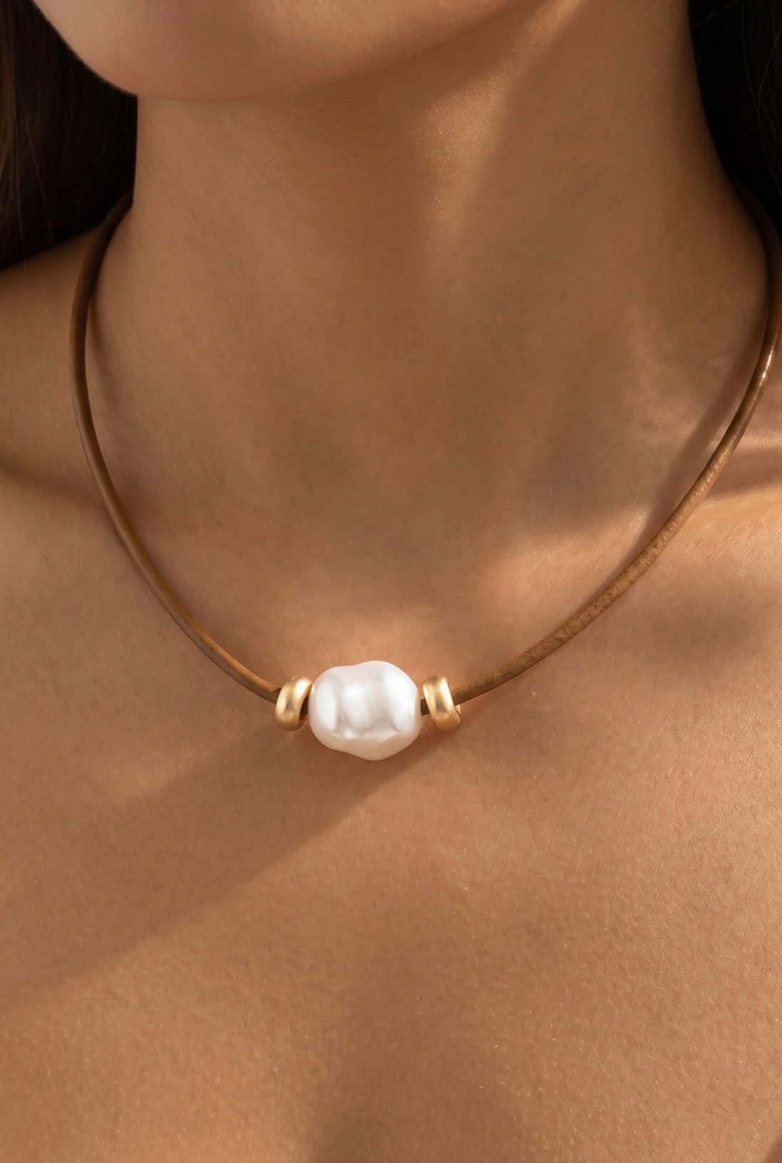 Brown leather cord necklace featuring an oversized baroque-style ceramic pearl with gold-tone spacer accents and an adjustable chain closure.
