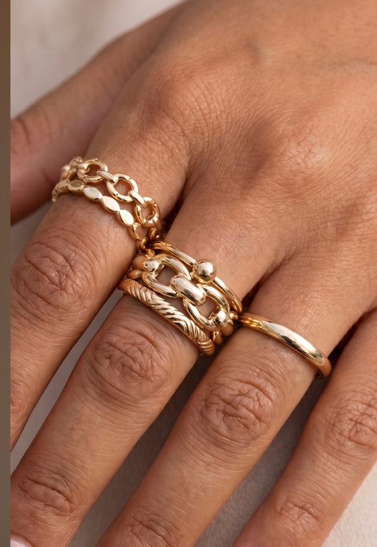 Five-piece gold ring stack set featuring engraved, polished dome, chain link, ball-and-bar, and Figaro oval link bands in a coordinated gold-tone finish.