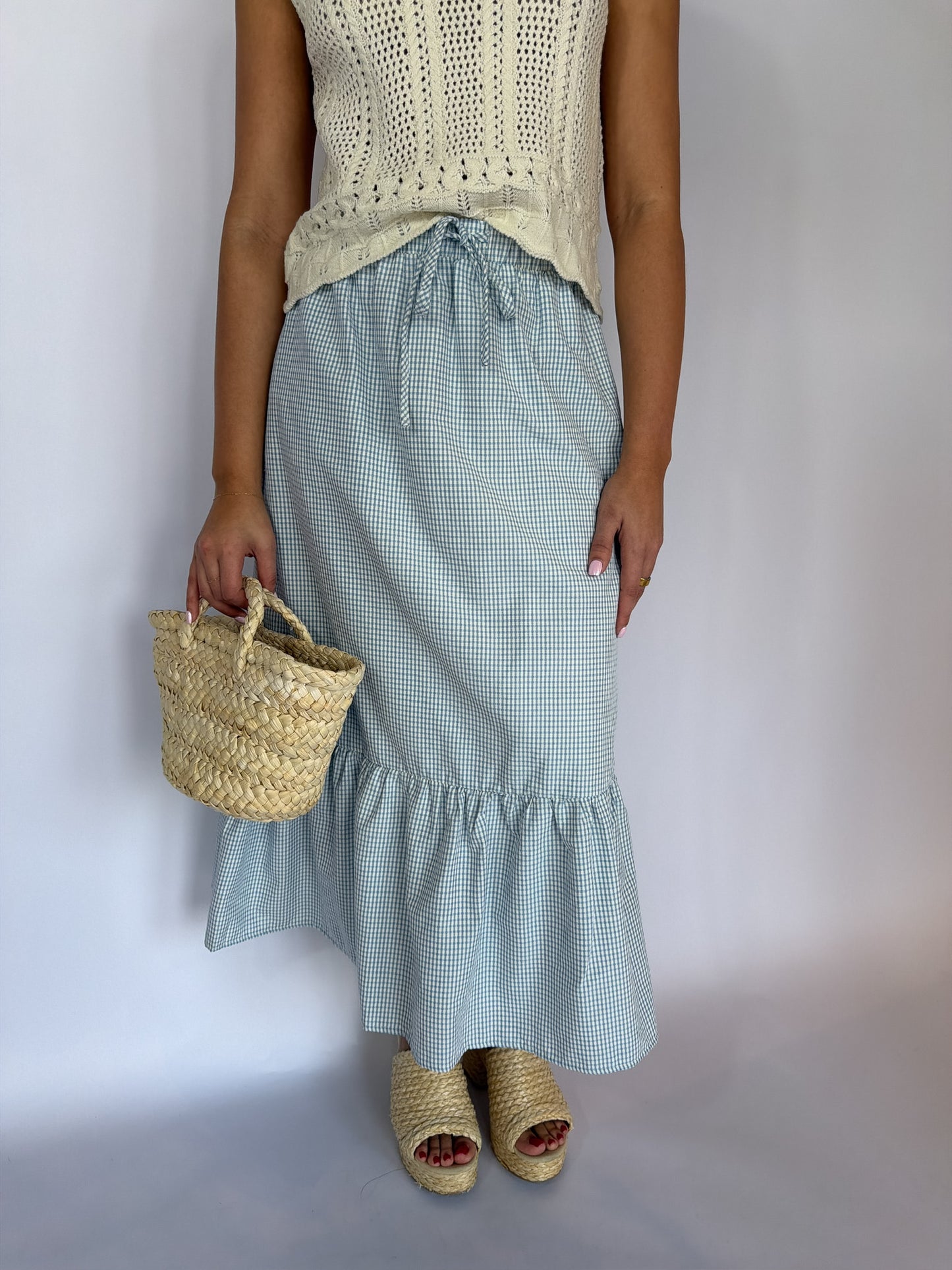 Person wearing a light blue checkered skirt and beige top, holding a straw bag on a plain background