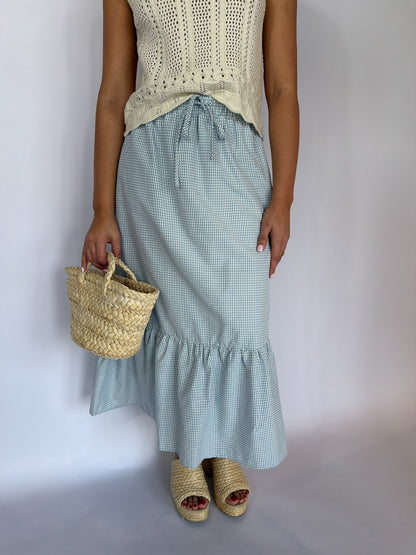 Person wearing a light blue checkered skirt and beige top, holding a straw bag on a plain background