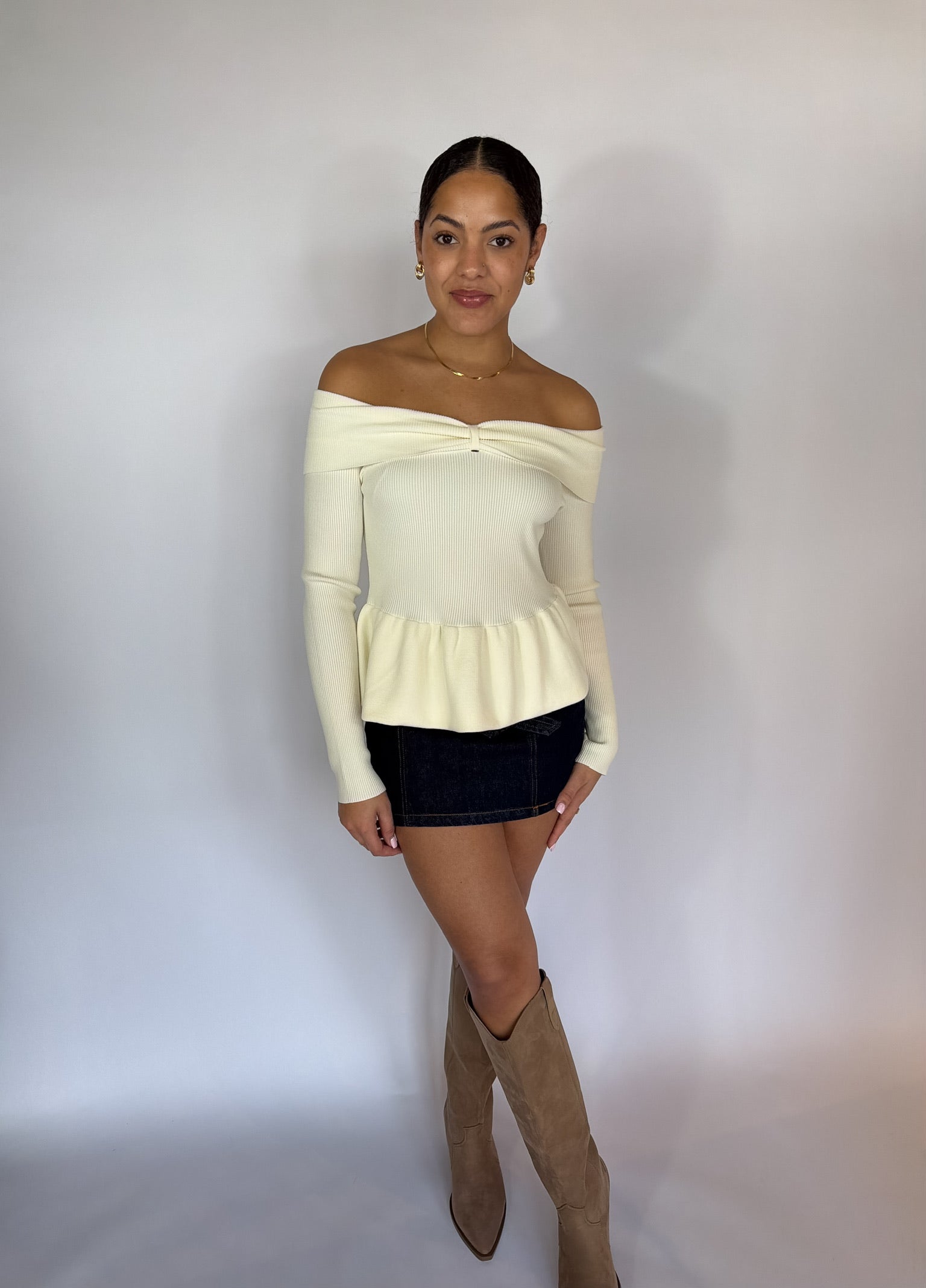 Woman wearing a cream off-shoulder sweater, black skirt, and brown knee-high boots against a plain background