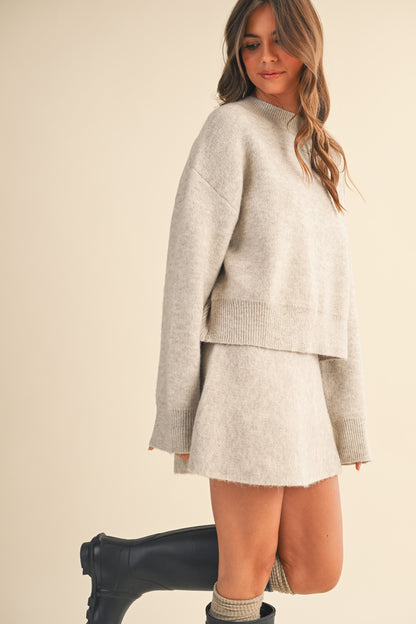 Woman wearing a matching knit sweater and skirt set with black rain boots on a beige background