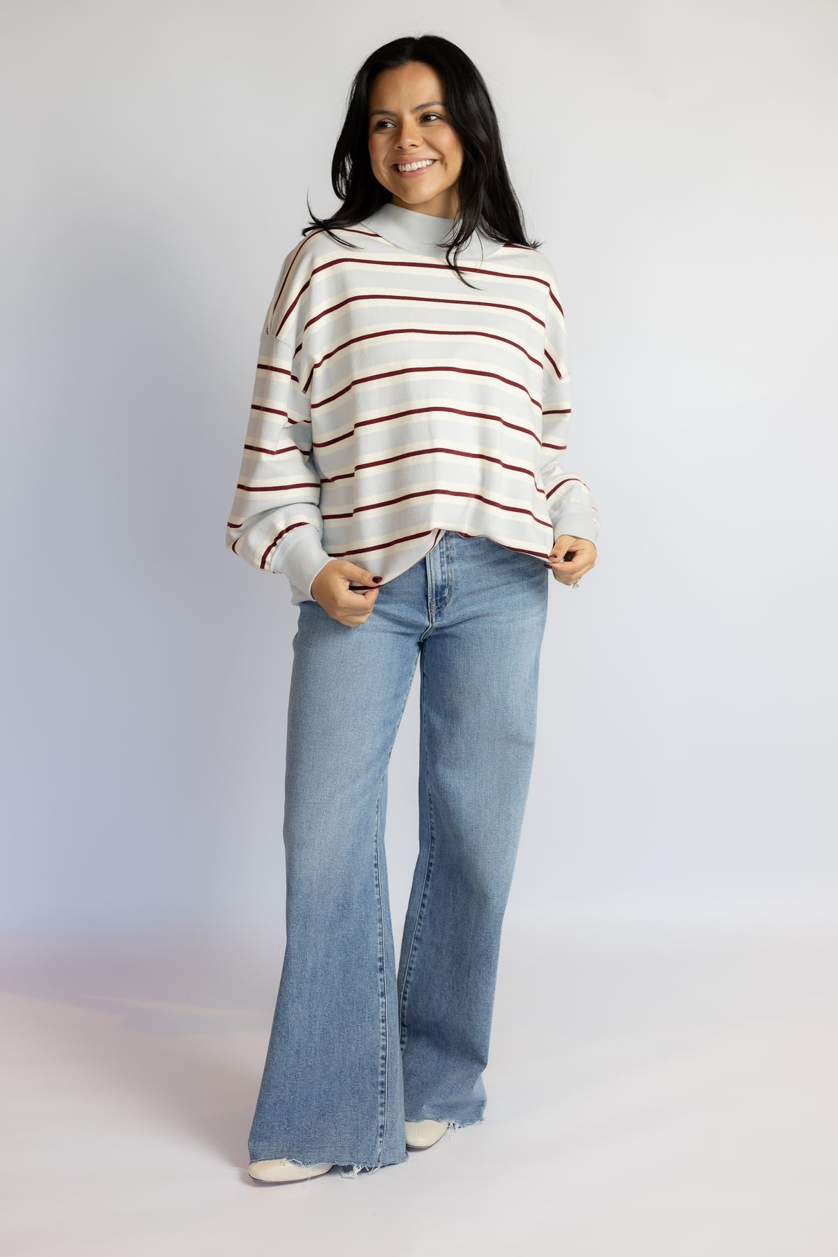 A woman modeling a cream knit sweater with thin red horizontal stripes and light-wash blue flare jeans, standing against a plain light gray studio background.