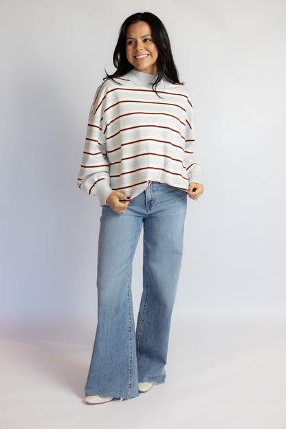 A woman modeling a cream knit sweater with thin red horizontal stripes and light-wash blue flare jeans, standing against a plain light gray studio background.