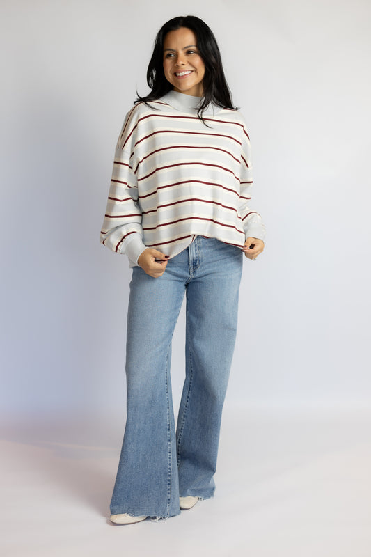 A woman modeling a cream knit sweater with thin red horizontal stripes and light-wash blue flare jeans, standing against a plain light gray studio background.