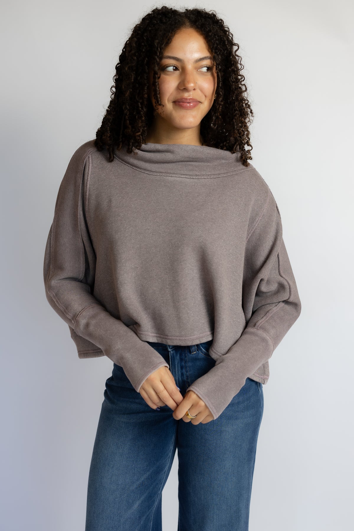 A woman with curly dark hair wearing a taupe-brown cowl-neck long-sleeve knit top and blue jeans, standing against a light gray studio background.