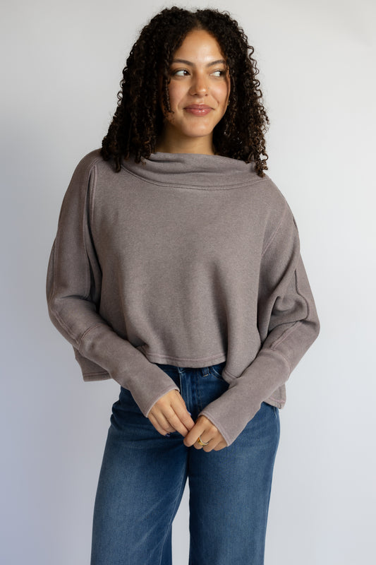 A woman with curly dark hair wearing a taupe-brown cowl-neck long-sleeve knit top and blue jeans, standing against a light gray studio background.