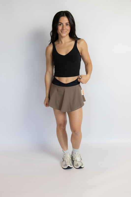 A woman modeling a black ribbed tank top and a taupe-brown athletic skirt, standing against a light gray studio background.