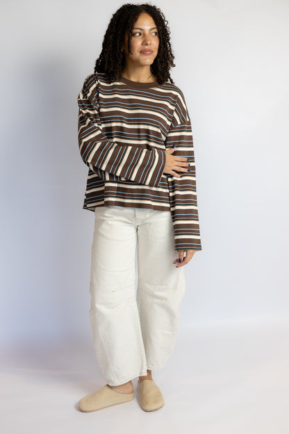 A woman modeling a brown, beige, and white striped long-sleeve top with voluminous balloon sleeves, paired with white wide-leg jeans and beige slip-on shoes, against a light gray studio background.