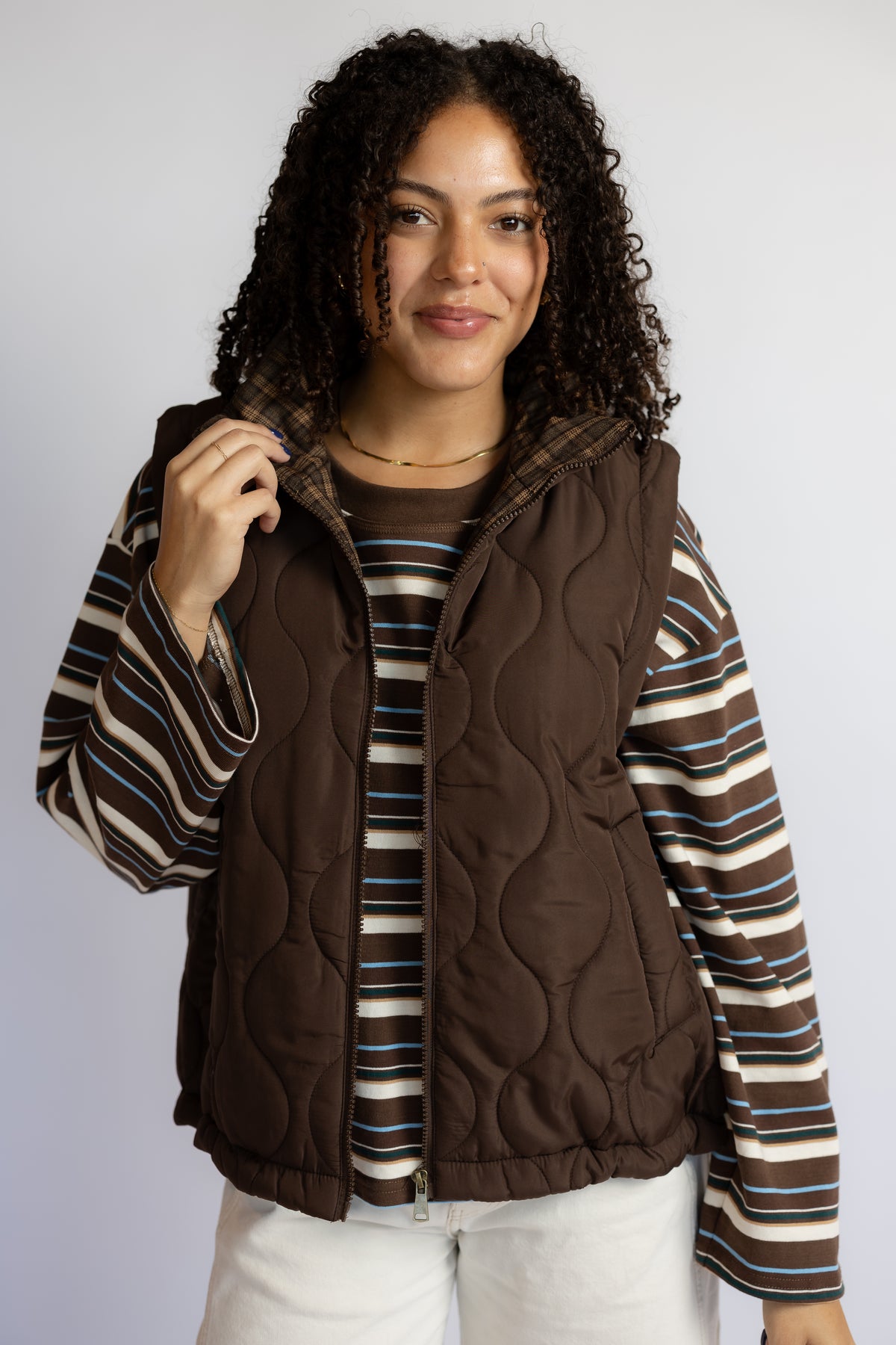 A woman wearing a brown quilted puffer vest over a brown, beige, and blue striped long-sleeve shirt, paired with white pants, against a light gray studio background.
