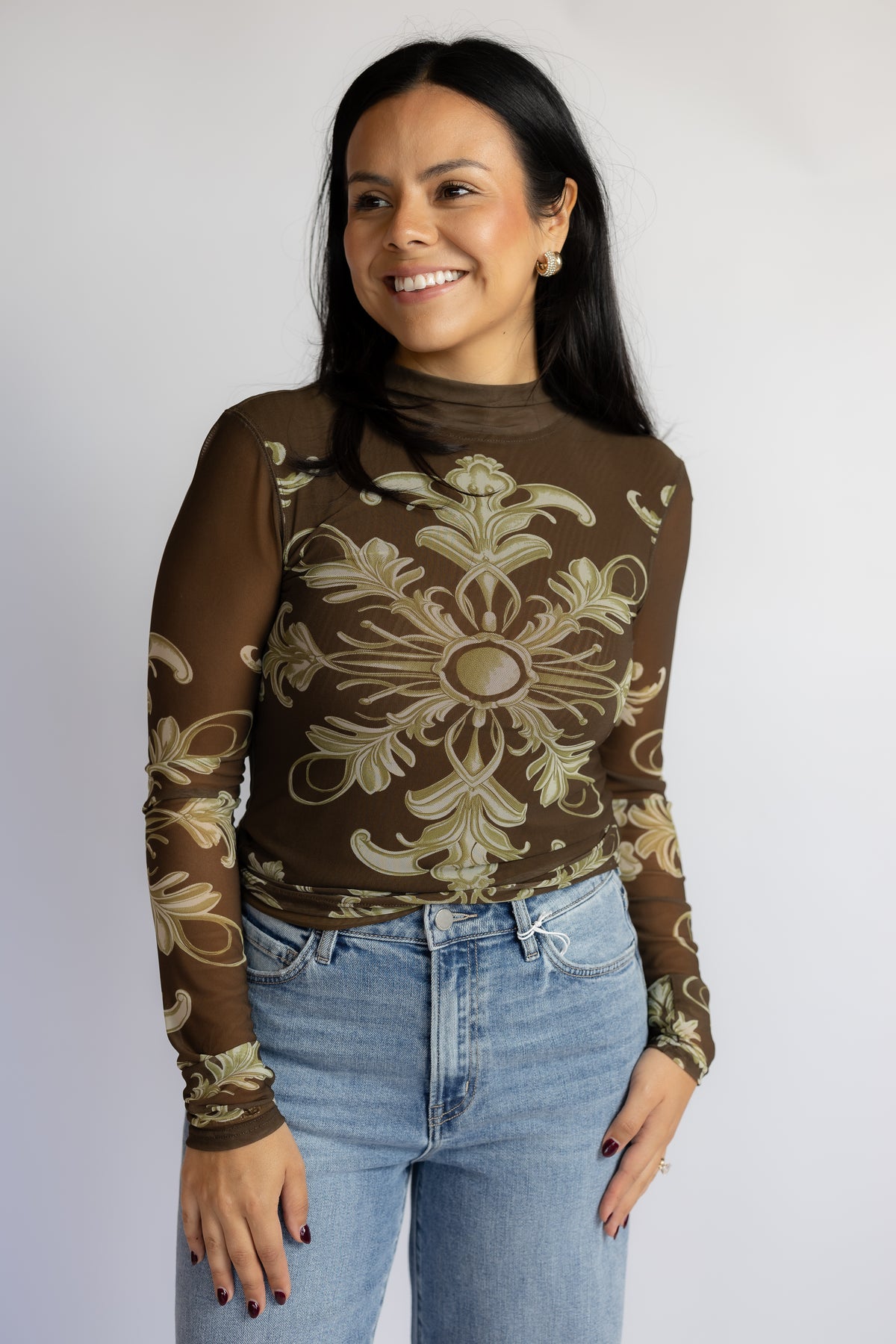 A woman wearing a brown mock-neck long-sleeve top with a beige baroque-style ornamental pattern, paired with light-wash blue jeans, standing against a light gray studio background.