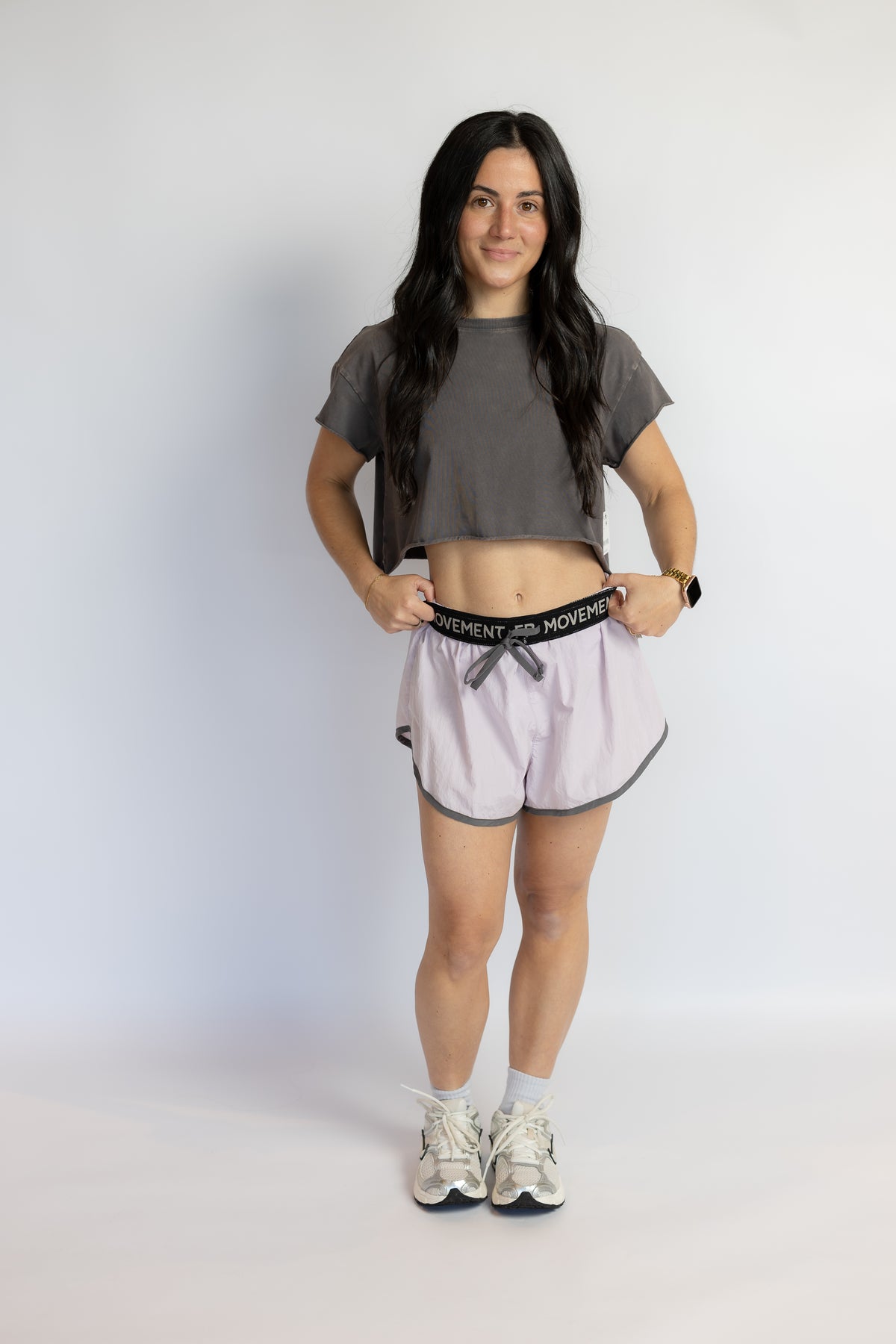 A woman modeling a pair of light pink athletic shorts with a black waistband that reads 'FP MOVEMENT', paired with a dark gray crop top and white sneakers, standing against a plain white studio background.
