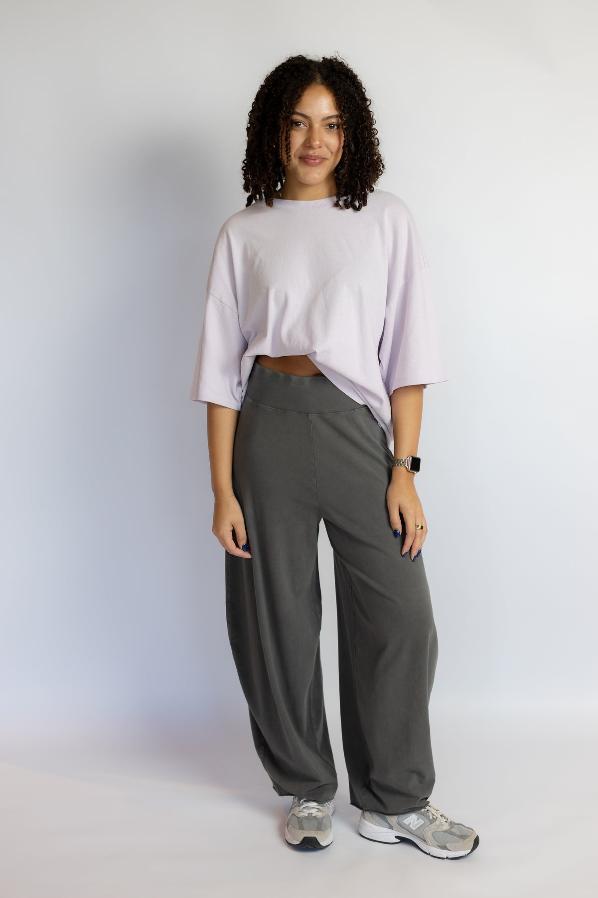 A woman modeling a light lavender oversized crop top and dark gray wide-leg sweatpants, standing against a plain white studio background.