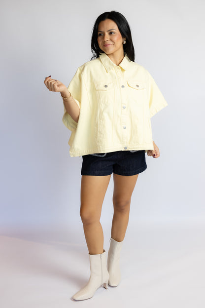 A woman modeling a pale yellow short-sleeve button-up shirt jacket, paired with dark denim shorts and off-white ankle boots, against a light gray studio background.
