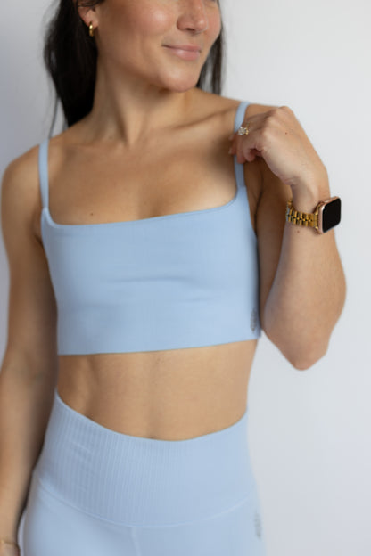 A woman wearing a light blue athletic crop top and matching high-waisted leggings, shown from the chest down against a plain light gray background.
