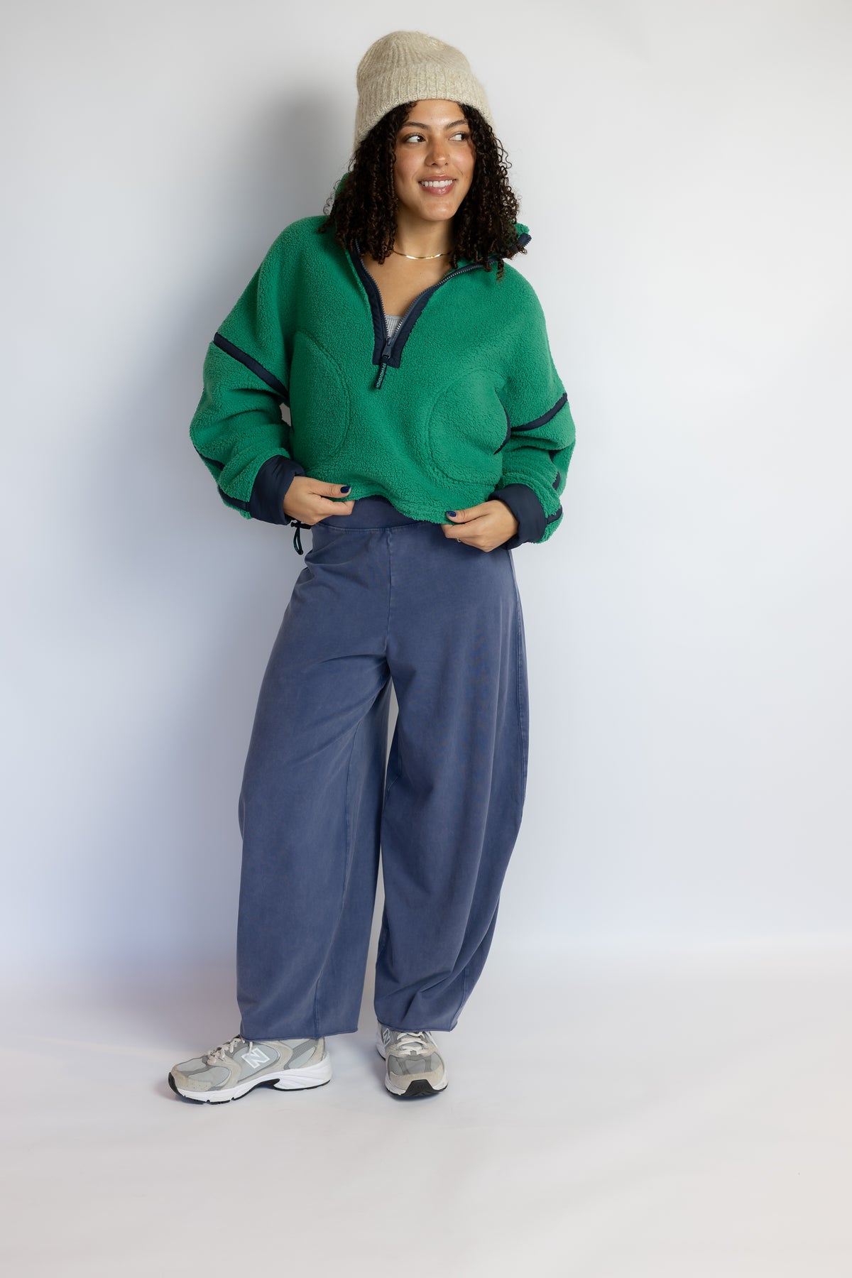 A woman modeling a green half-zip fleece pullover with dark trim, paired with wide-leg blue sweatpants, a beige beanie, and gray sneakers, standing against a plain light gray studio background.