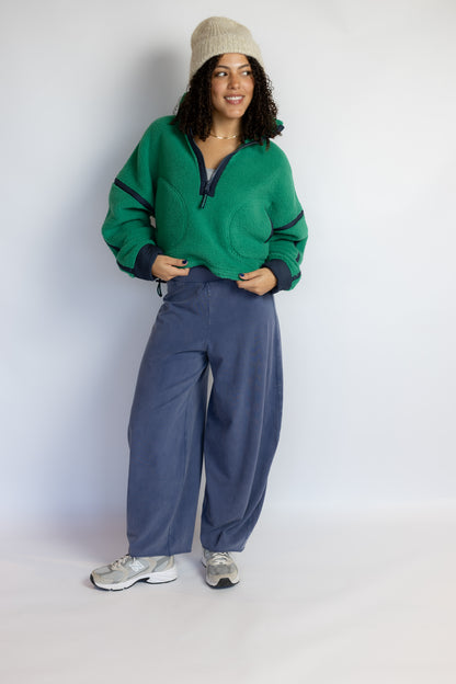 A woman modeling a green half-zip fleece pullover with dark trim, paired with wide-leg blue sweatpants, a beige beanie, and gray sneakers, standing against a plain light gray studio background.