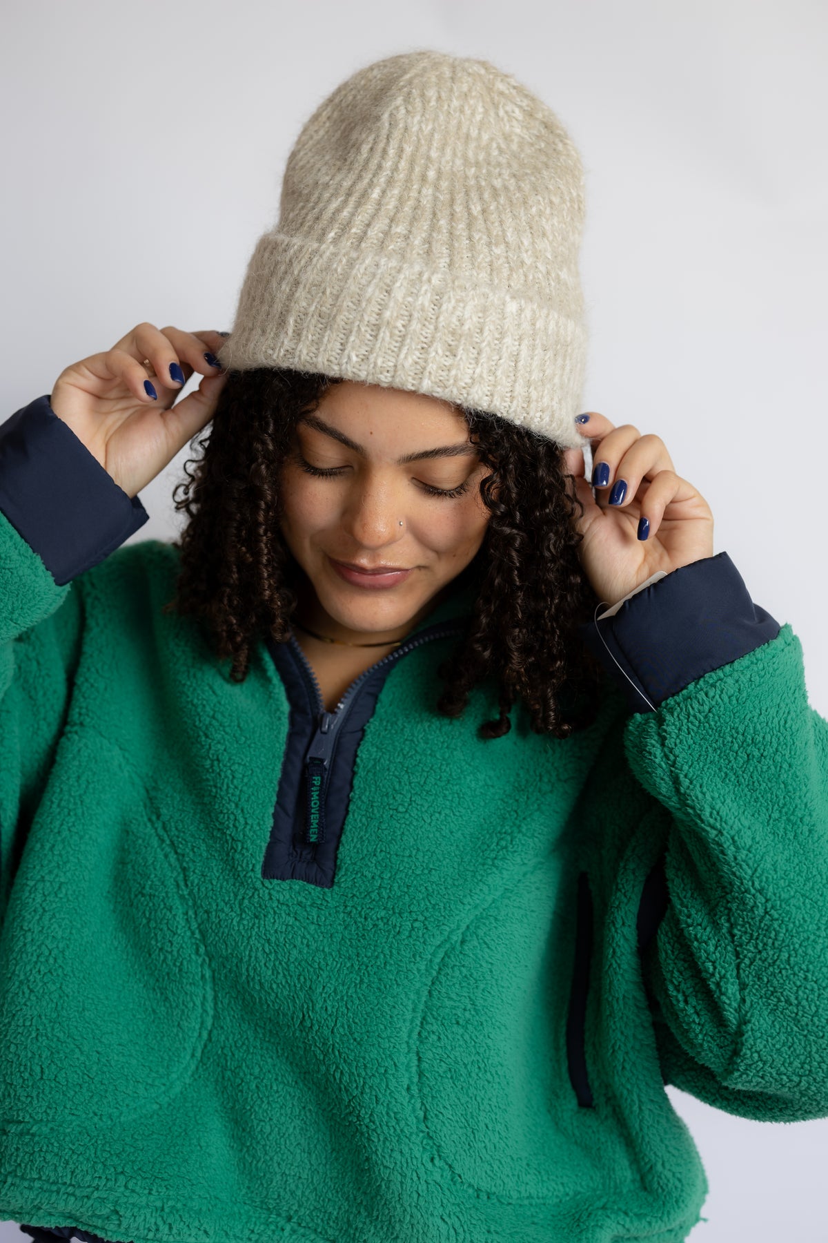 A woman wearing a cream-colored ribbed knit beanie and a green fleece pullover with dark navy trim, posing against a light gray background.