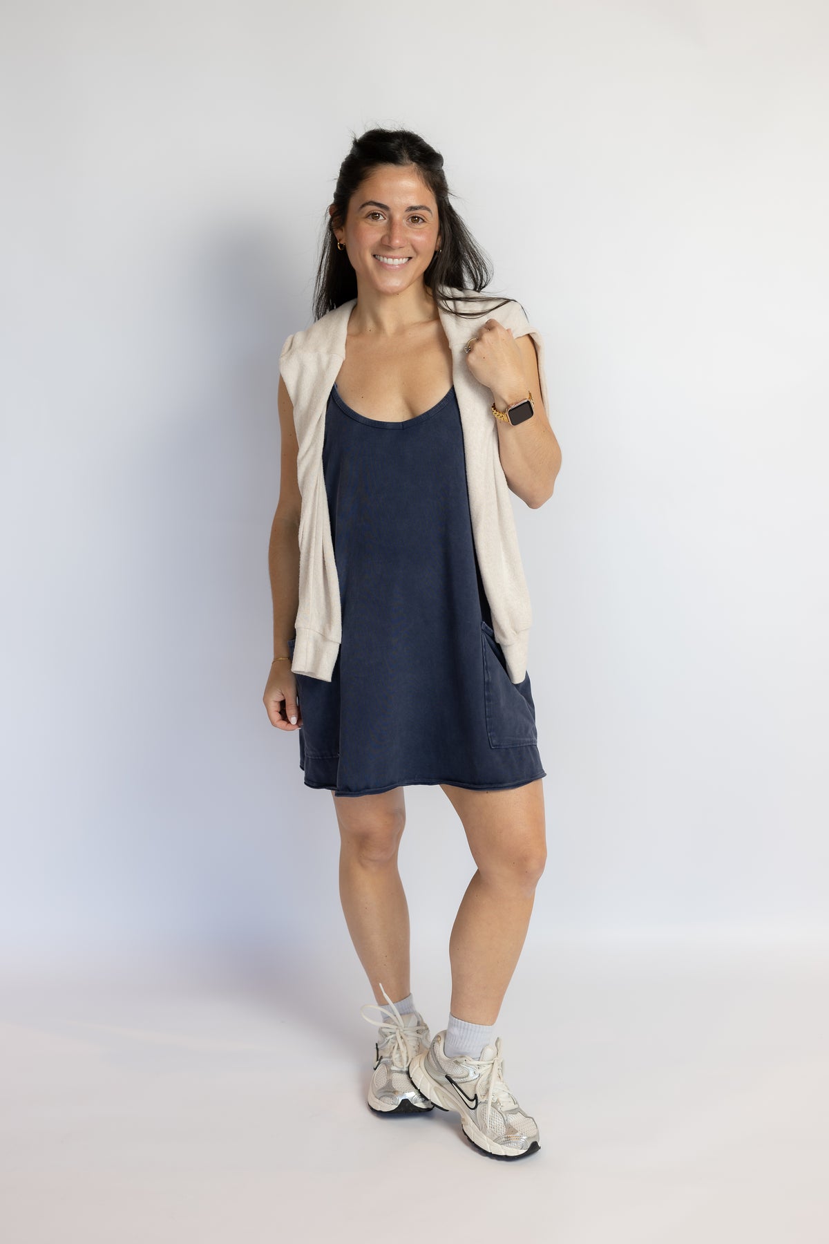 A woman modeling a sleeveless cream vest over a navy blue tankini dress, paired with white athletic sneakers, standing against a plain white studio background.