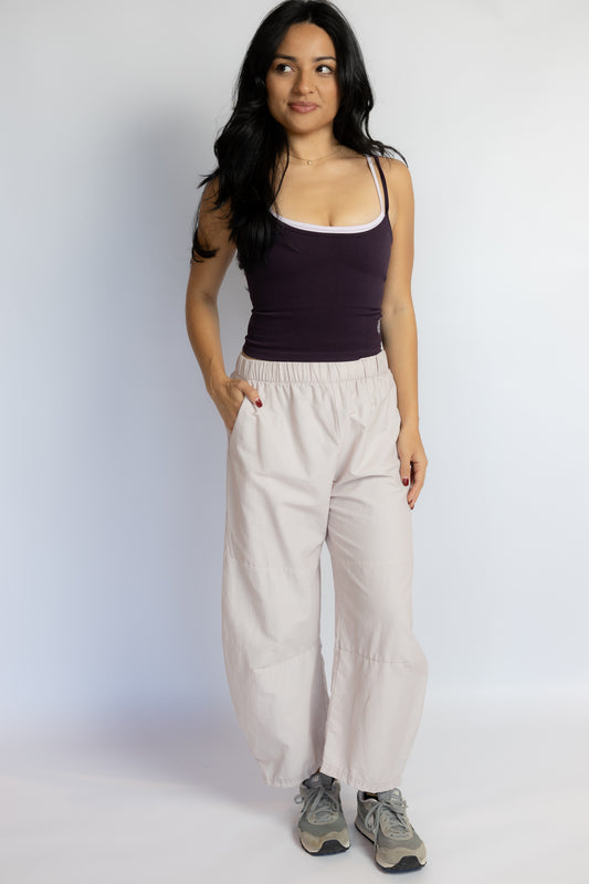 A woman modeling a dark plum tank top and light beige wide-leg pants with gray sneakers, standing against a light gray studio background.