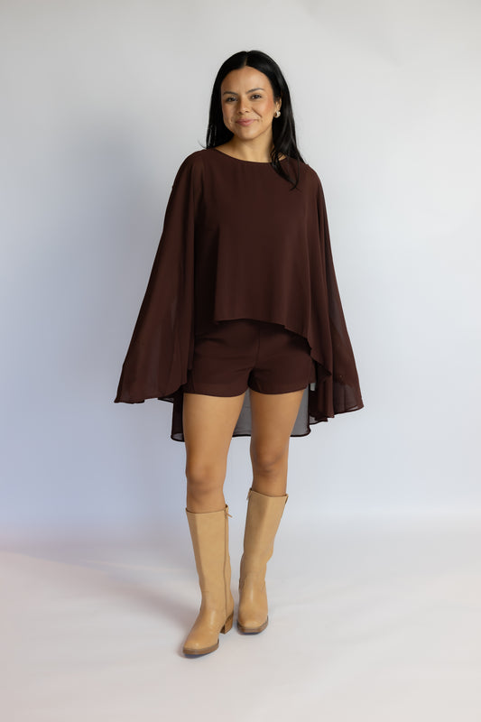 A woman modeling a dark brown two-piece outfit consisting of a long-sleeve draped poncho-style top and matching shorts, paired with tan knee-high boots, against a light gray studio background.
