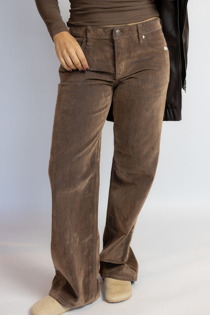 A person wearing brown wide-leg corduroy pants, styled with a brown long-sleeve top, a black jacket, and beige slip-on shoes against a light gray studio background.