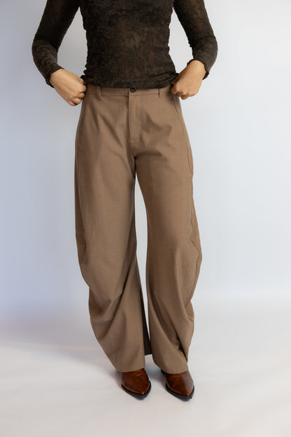 A person wearing loose, wide-leg taupe-brown trousers with a dark brown lace long-sleeve top and brown leather shoes, standing against a light gray studio background.