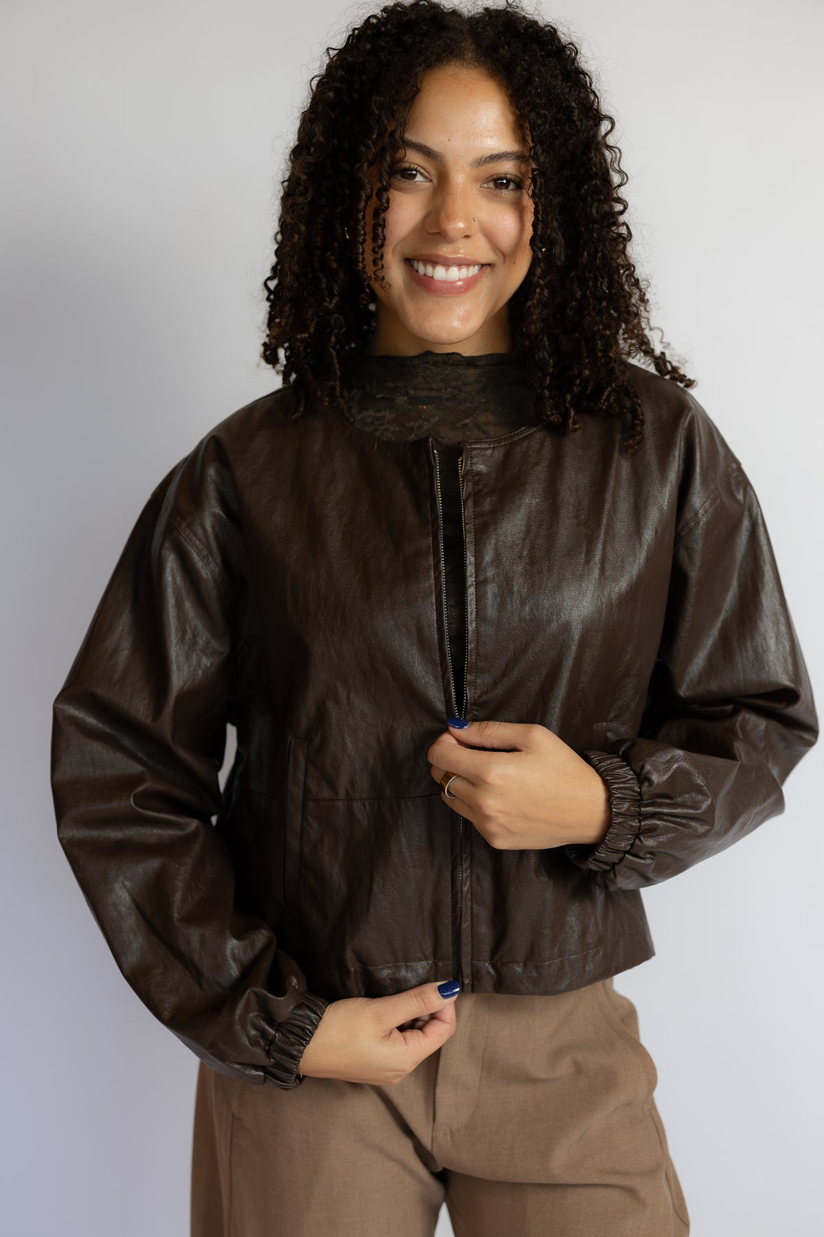A smiling woman with curly dark hair models a dark brown faux-leather jacket with a high mock neck and gathered cuffs, paired with brown pants, against a plain light gray background.