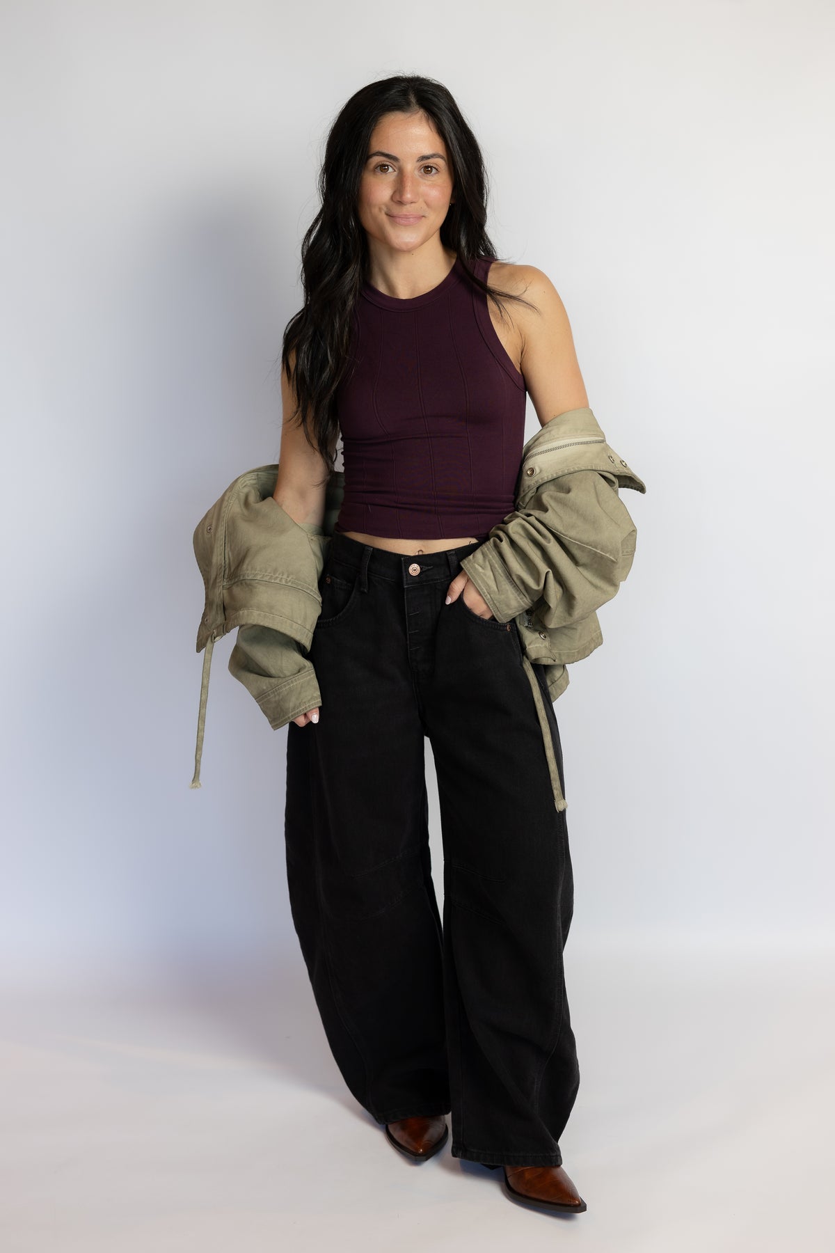 A woman modeling a dark plum ribbed tank top, black wide-leg pants, and an olive green jacket draped off her shoulders, standing against a light gray studio background.