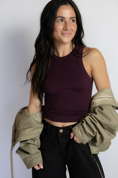 A woman modeling a ribbed sleeveless crop top in deep plum purple, styled with black jeans and an olive green jacket draped off her shoulders, against a plain light gray background.