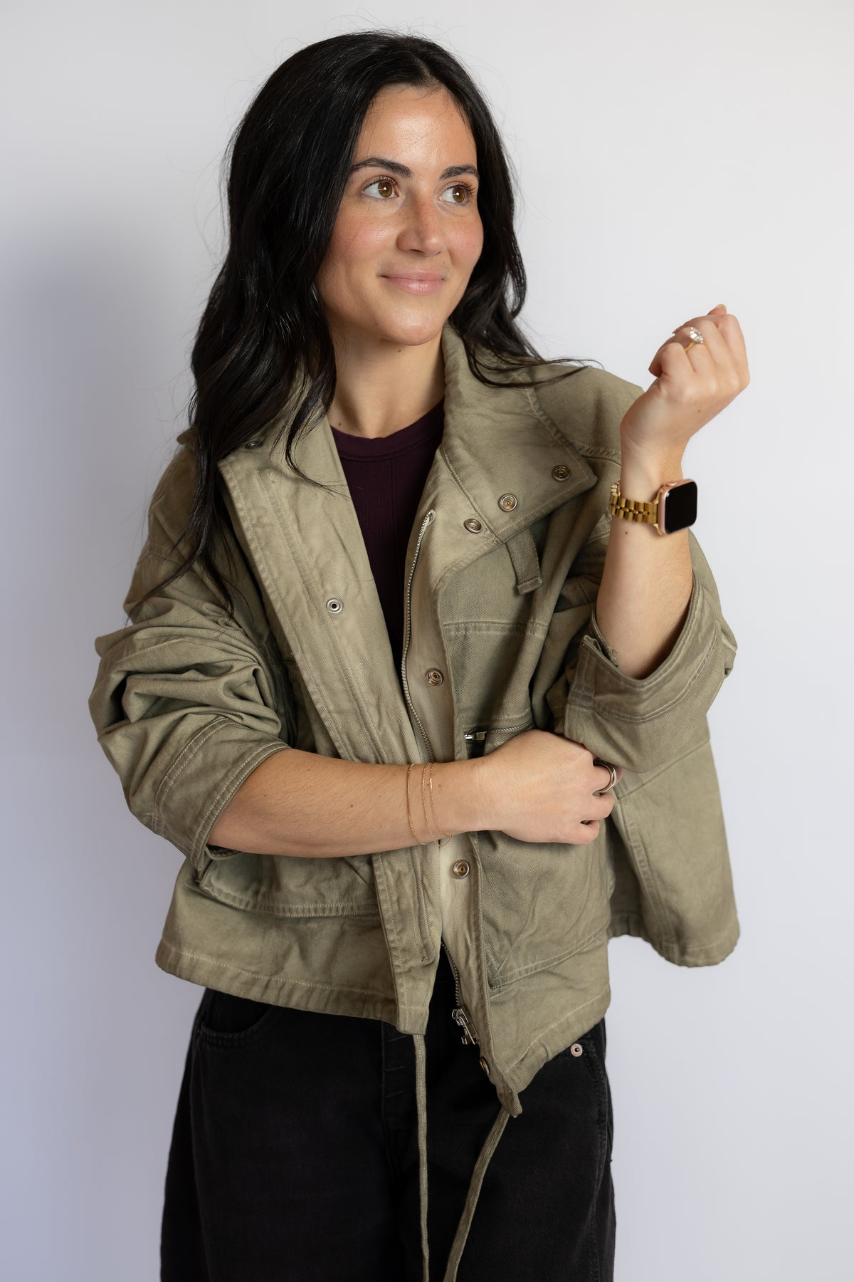 A woman wearing a cropped olive green utility jacket over a dark plum top and black pants, posing against a light gray studio background.