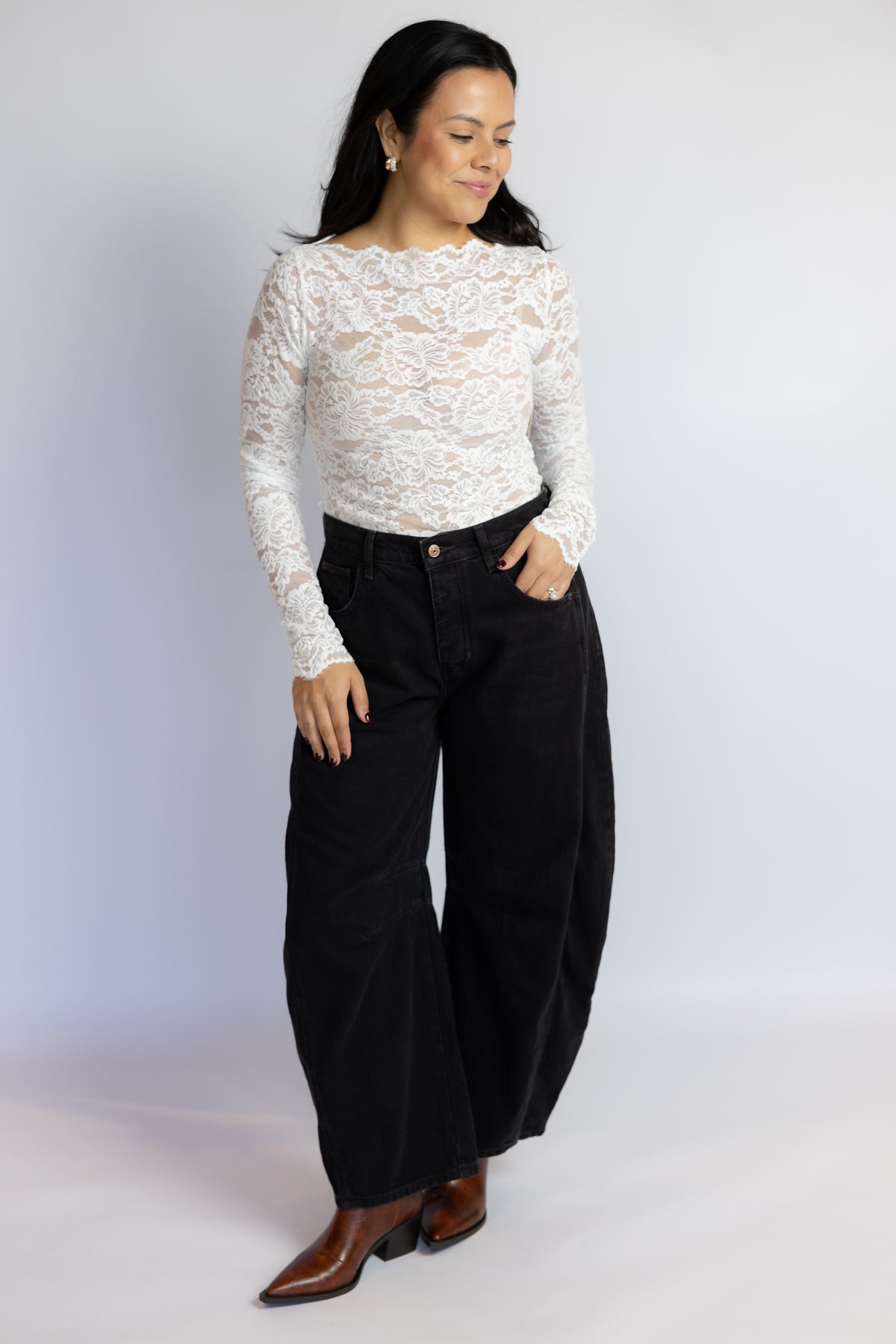 A woman modeling a white long-sleeve lace top paired with black wide-leg pants and brown pointed-toe heeled boots, standing against a light gray studio background.