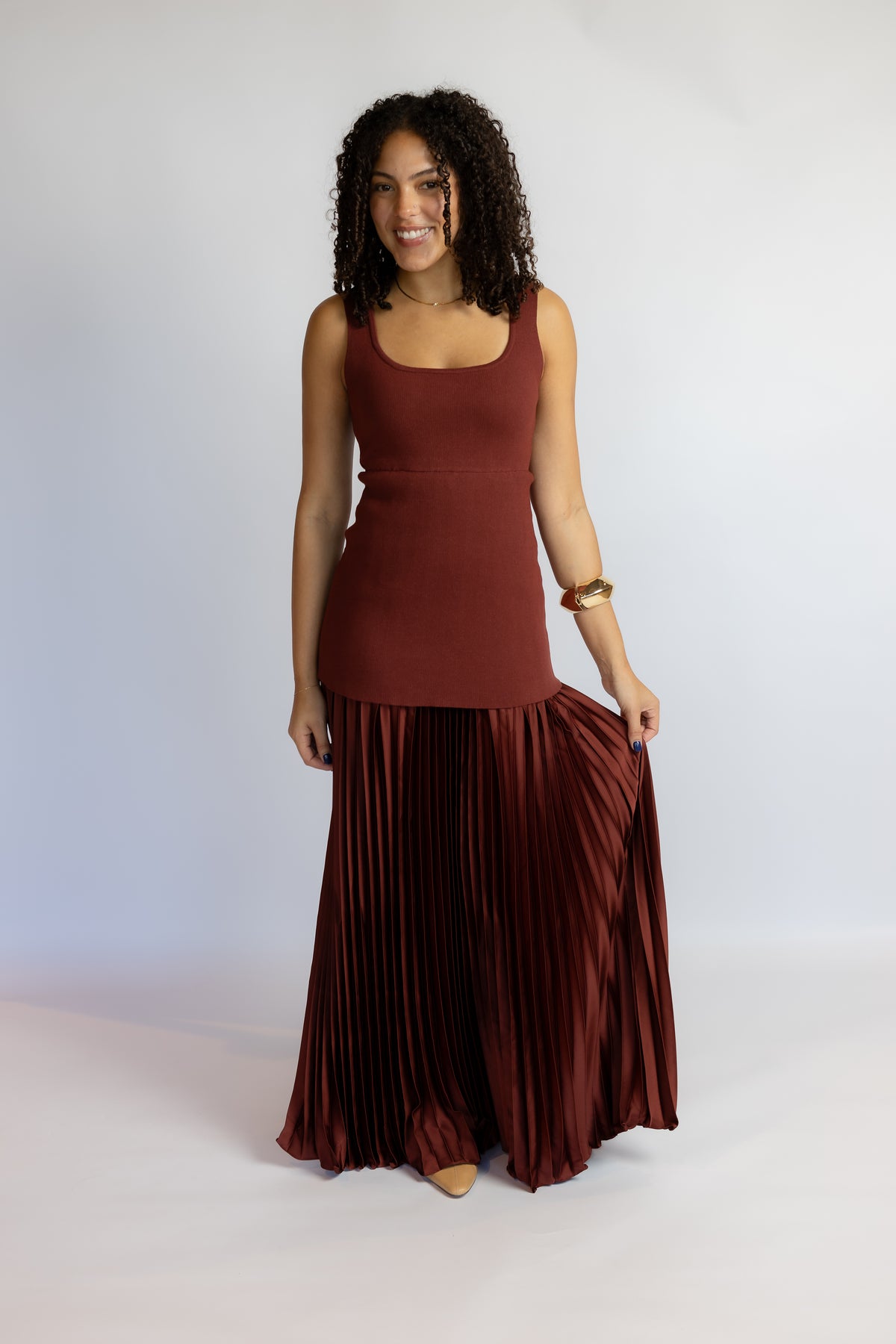 A woman modeling a sleeveless rust-brown top and matching pleated maxi skirt against a plain light gray studio background.