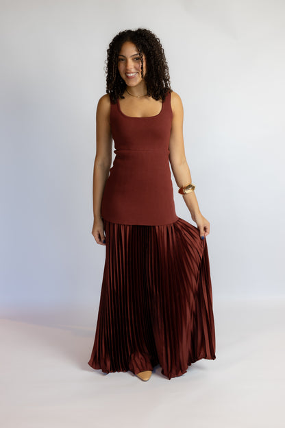 A woman modeling a sleeveless rust-brown top and matching pleated maxi skirt against a plain light gray studio background.