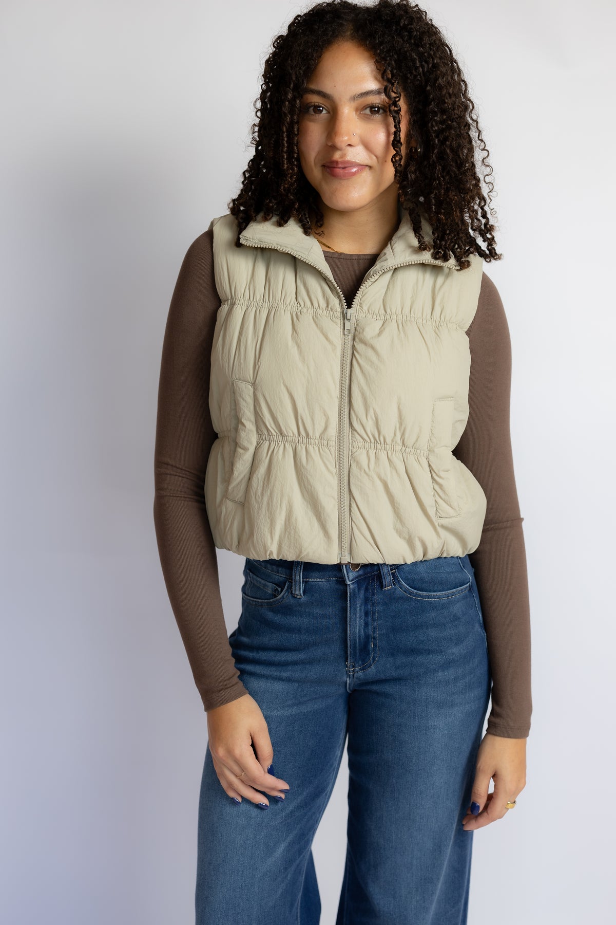 A woman modeling a sleeveless quilted puffer vest in light beige, worn over a brown long-sleeve top and blue jeans, against a plain light gray studio background.