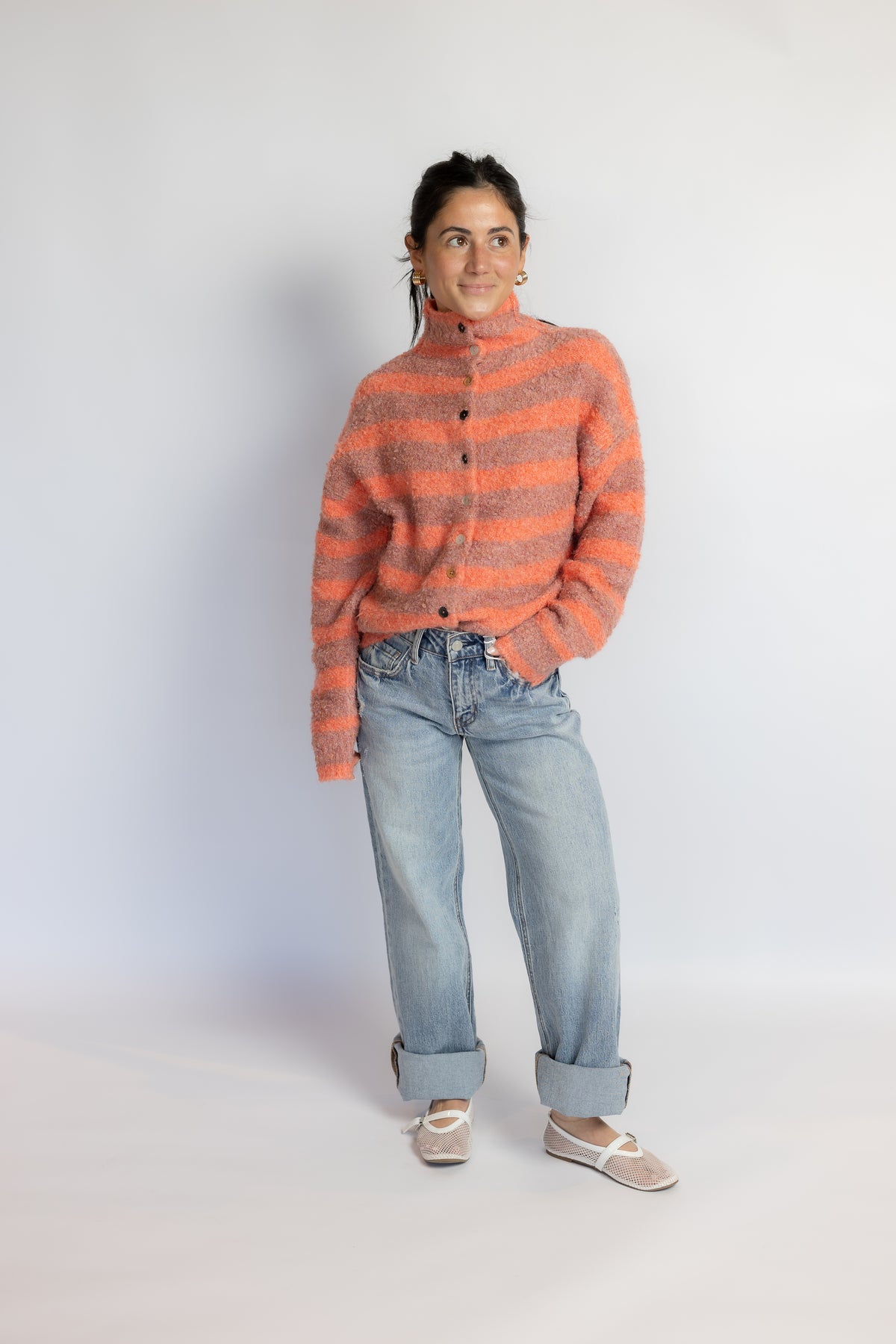 A woman stands against a plain light gray studio background wearing a fuzzy orange and gray striped mock-neck cardigan, light-wash blue jeans, and beige ballet flats.
